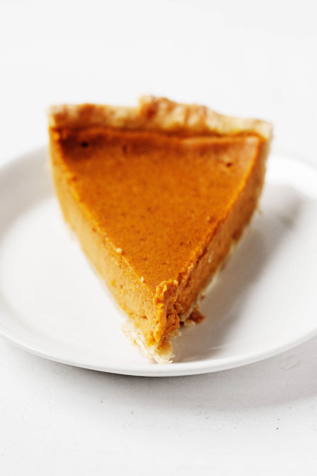 A single slice of pumpkin pie is resting on a small dessert plate on a white surface.