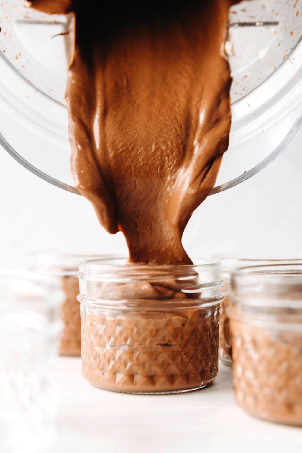 A smooth, blended chocolate pudding mixture is being poured into several small, glass jars.