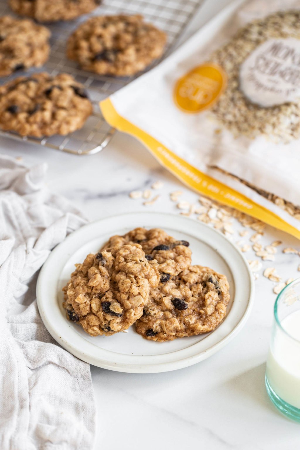 A few vegan oatmeal raisin cookies are stacked on a small white serving plate. A bag of rolled oats lies in the backdrop, next to a wire cooling rack.