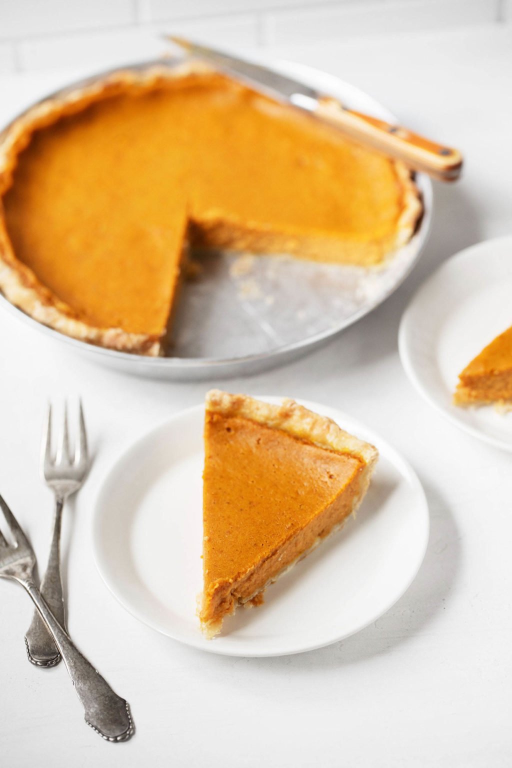 A silver pie pan holds a classic vegan pumpkin pie. A slice of the pie peeks out on a serving plate in front of it, with forks nearby.