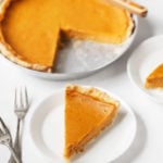 A silver pie pan holds a classic vegan pumpkin pie. A slice of the pie peeks out on a serving plate in front of it, with forks nearby.