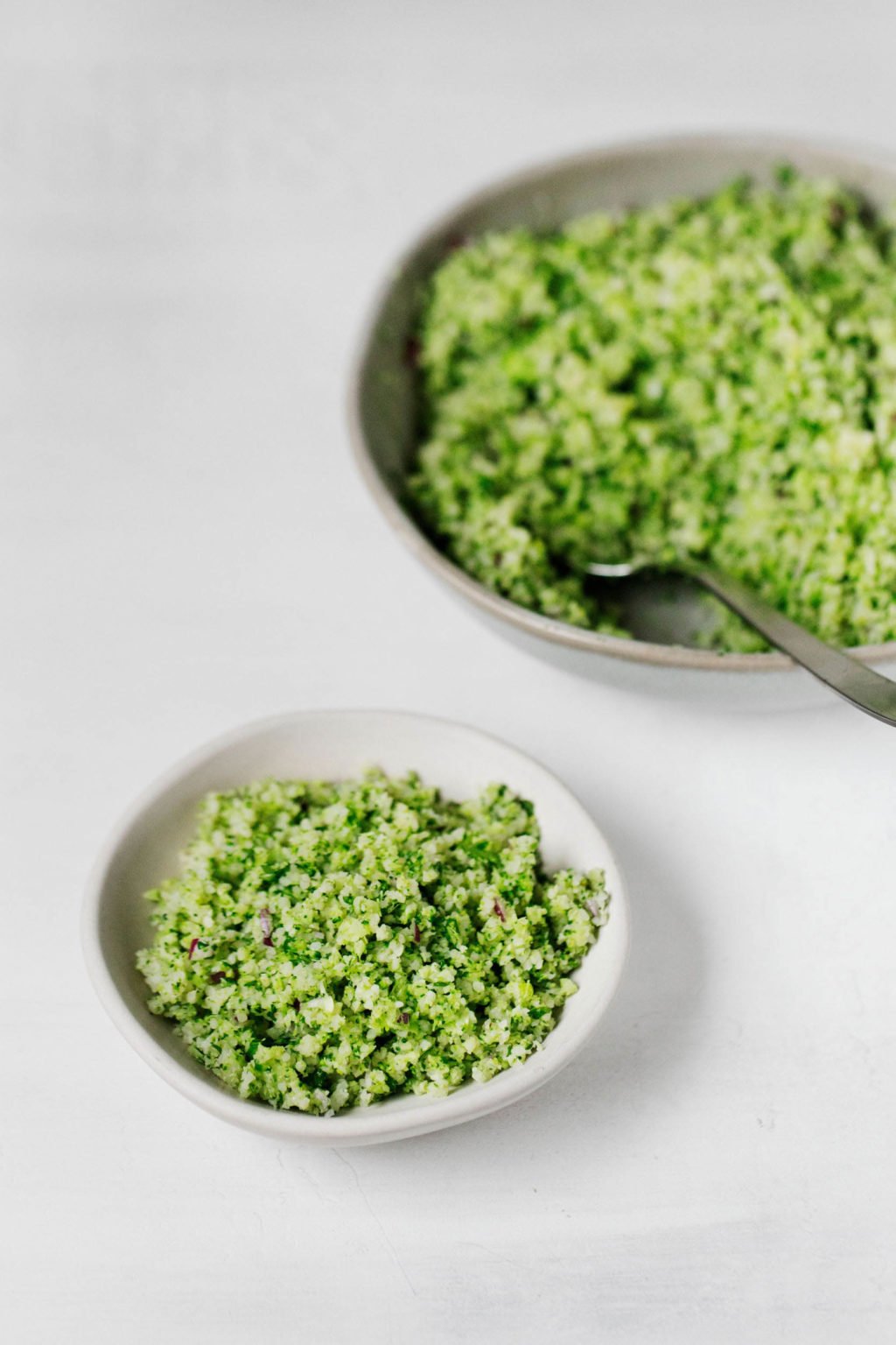 A skillet with cooked, chopped broccoli rests in the background. A white dish with a vegan broccoli tabbouleh is visible in front.