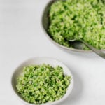 A skillet with cooked, chopped broccoli rests in the background. A white dish with a vegan broccoli tabbouleh is visible in front.