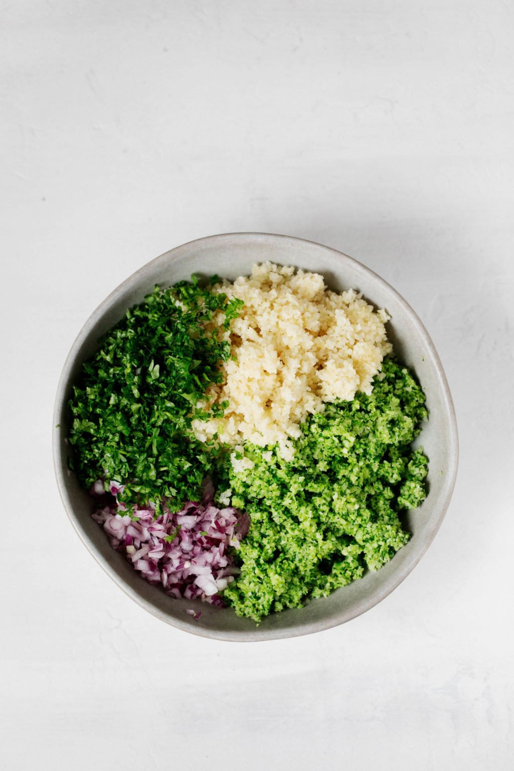 A mixing bowl contains the components for a vibrant, plant-based whole grain salad. The bowl rests on a white surface.