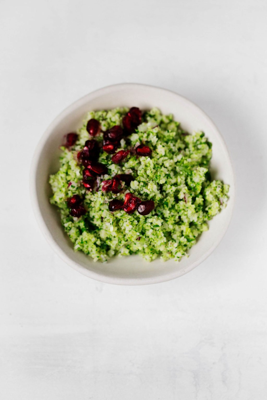 A small, white bowl of broccoli tabbouleh rests on a white surface. It's topped with pomegranate arils. 