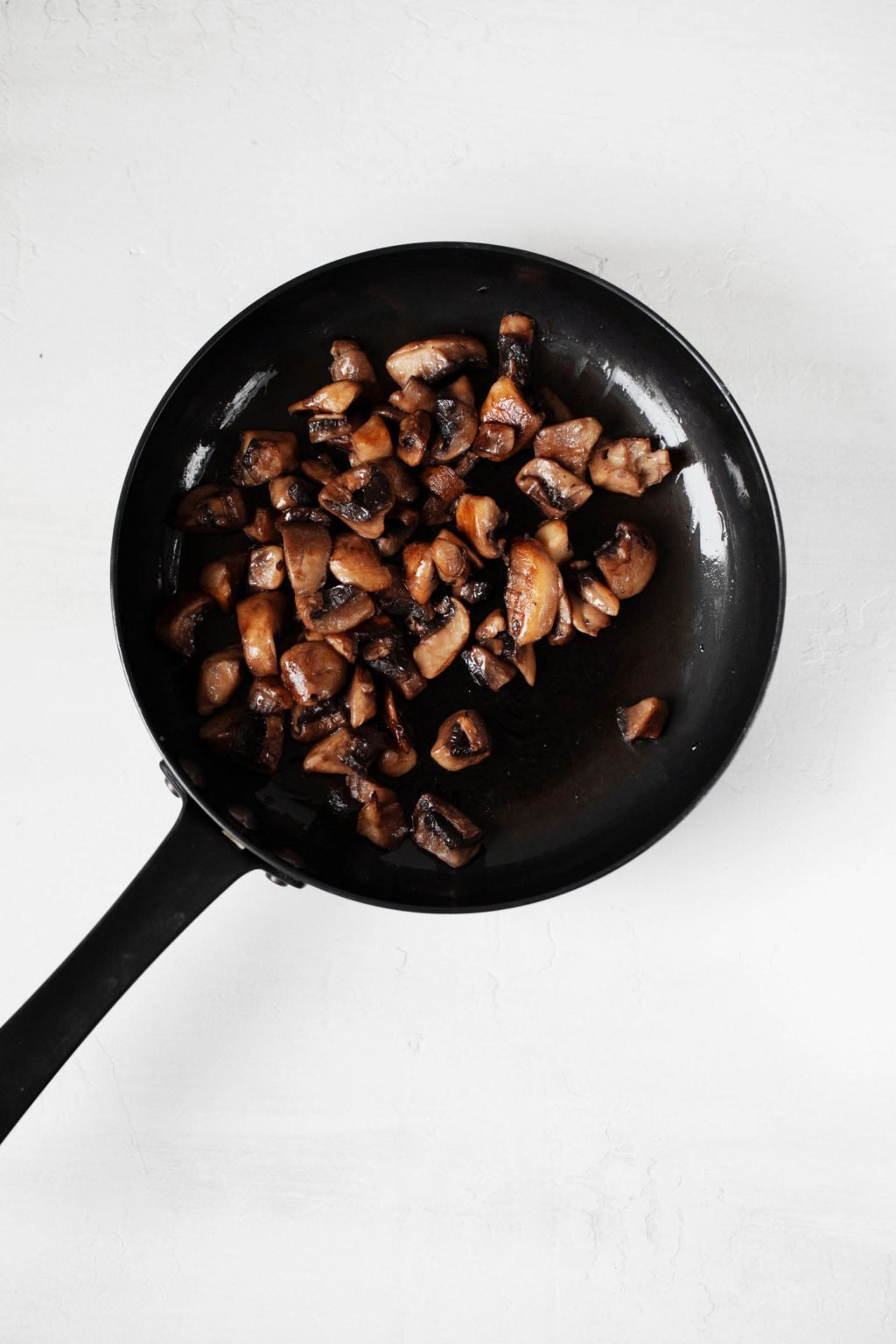 Sauteed mushrooms are pictured in a small, black frying pan. The pan rests on a white surface.