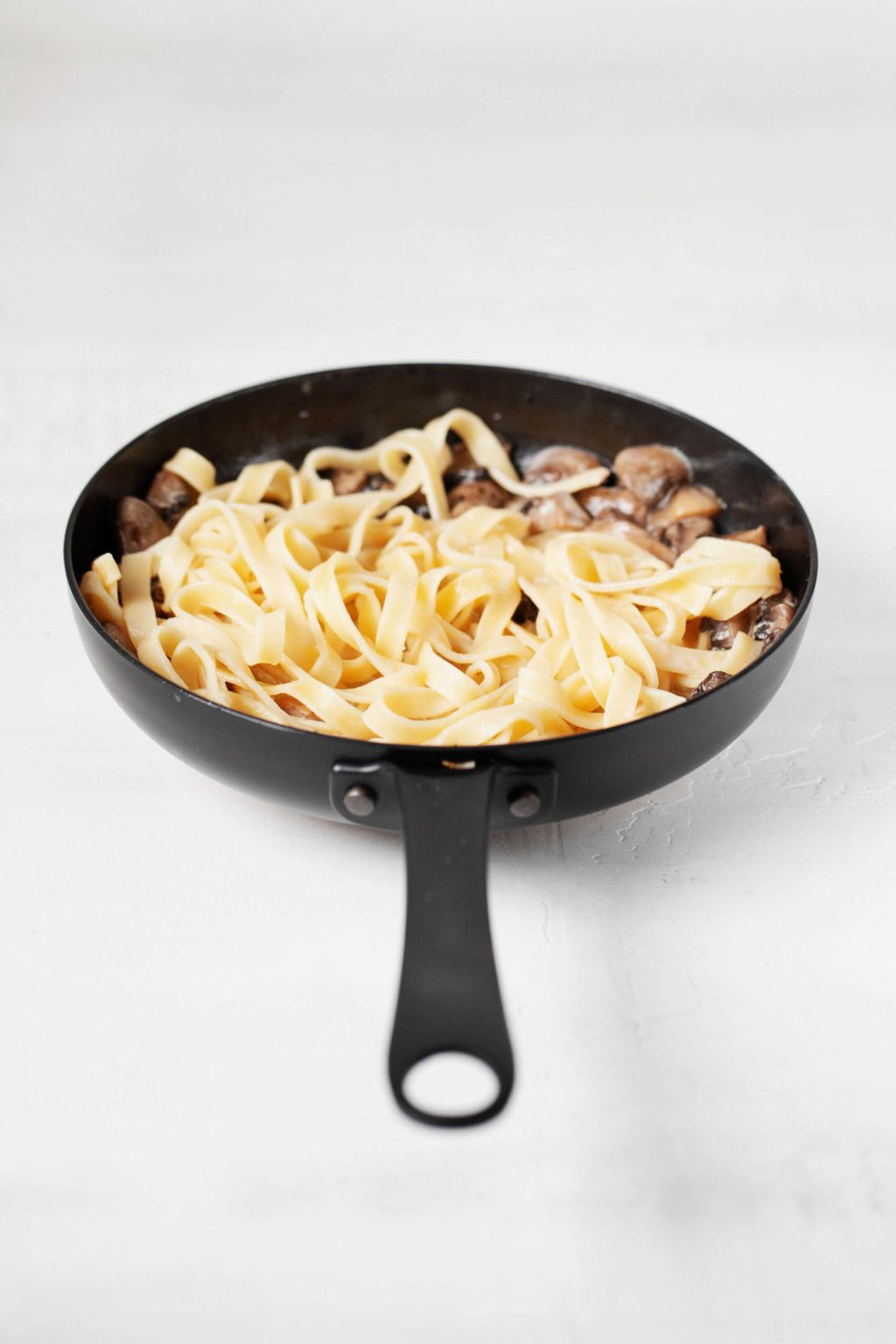A black, cast iron skillet is filled with fettucine, vegetables, and a creamy sauce.