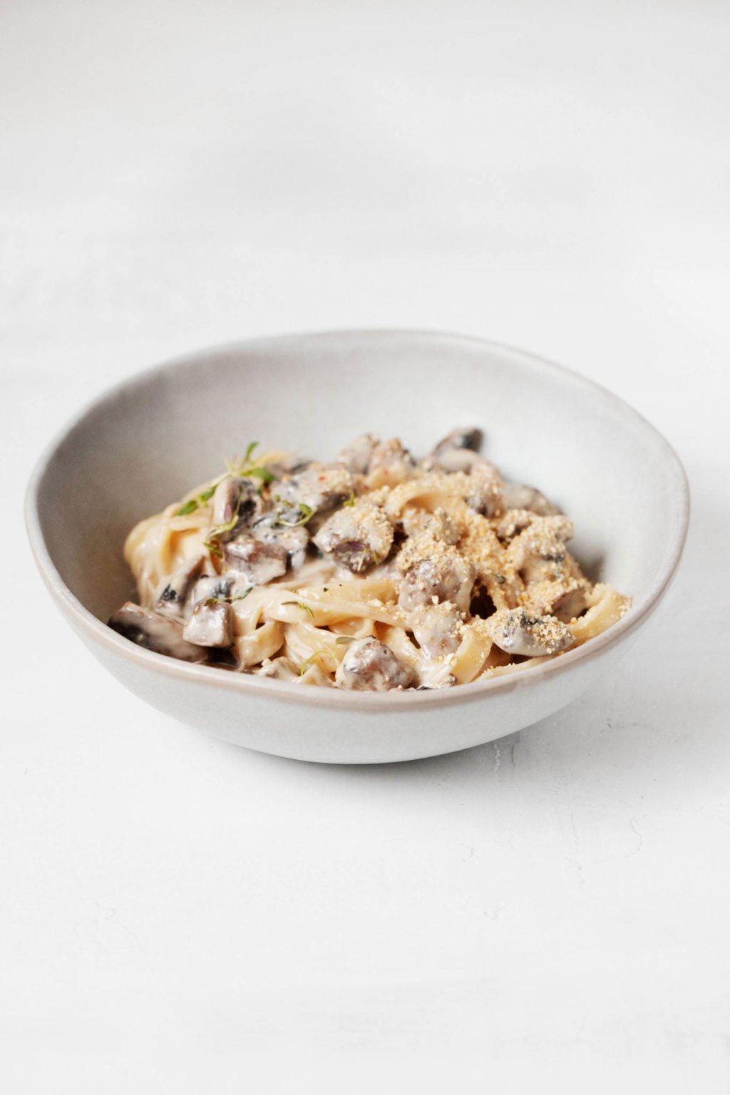 Een gekanteld beeld van een witte kom romige veganistische pasta met champignons, rustend op een witte ondergrond.