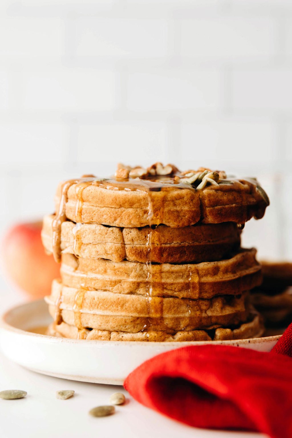 A stack of vegan apple cinnamon waffles is covered in pumpkin seeds and maple syrup. There's a red napkin nearby.