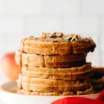 A stack of vegan apple cinnamon waffles is covered in pumpkin seeds and maple syrup. There's a red napkin nearby.