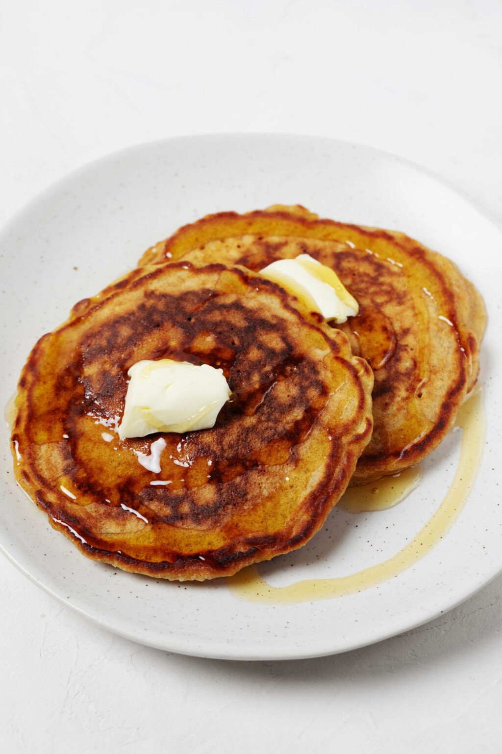 An image of two nicely browned vegan carrot cake pancakes, topped with a pat of butter and served on a round white plate.