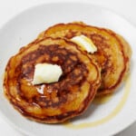 An image of two nicely browned vegan carrot cake pancakes, topped with a pat of butter and served on a round white plate.