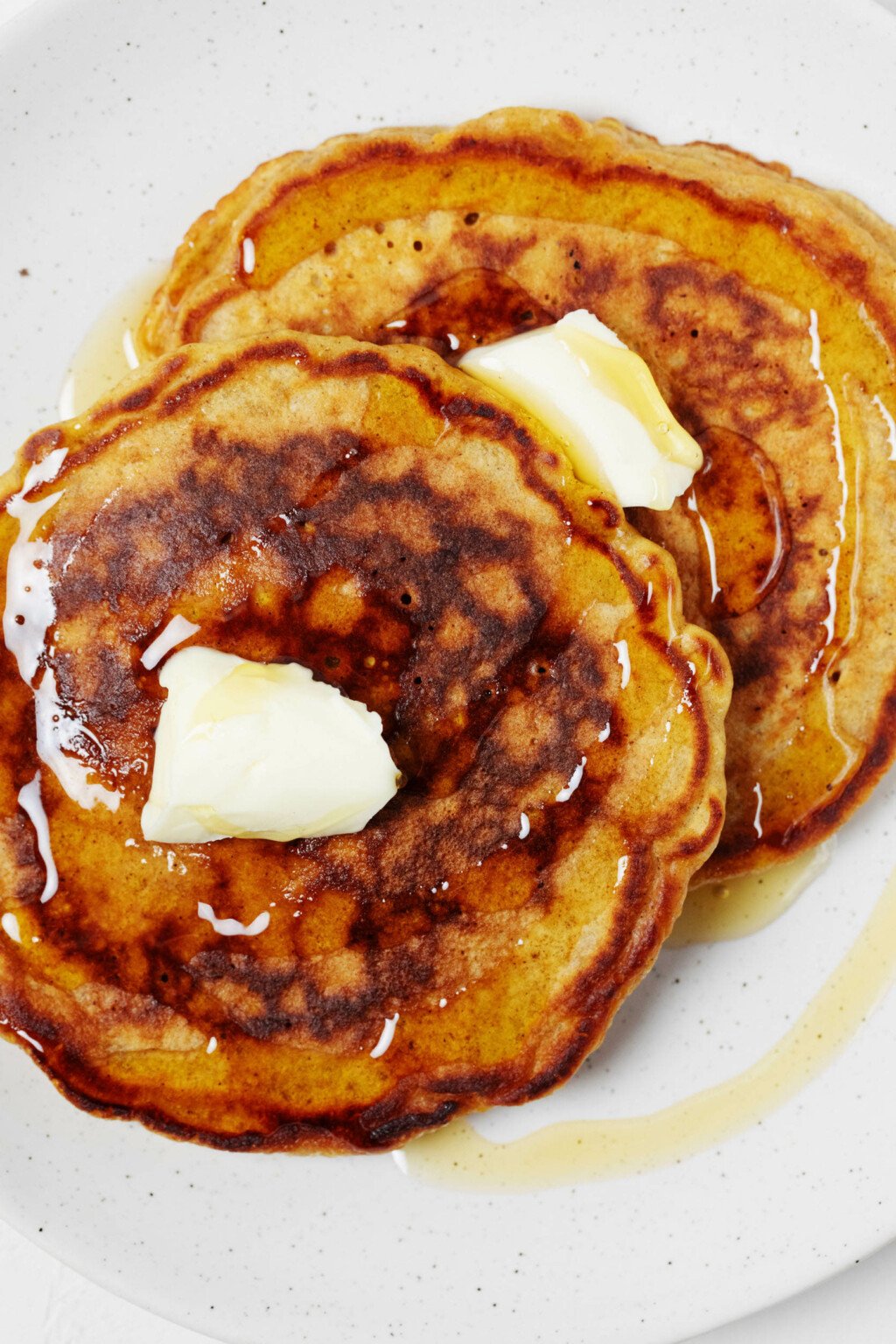 An image of two nicely browned vegan carrot cake pancakes, topped with a pat of butter and served on a round white plate.