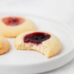 Three vegan thumbprint cookies with different colored jam centers are resting on a small, white dessert plate.