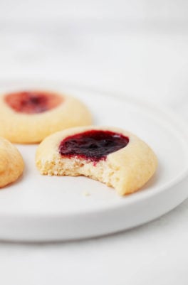 Three vegan thumbprint cookies with different colored jam centers are resting on a small, white dessert plate.