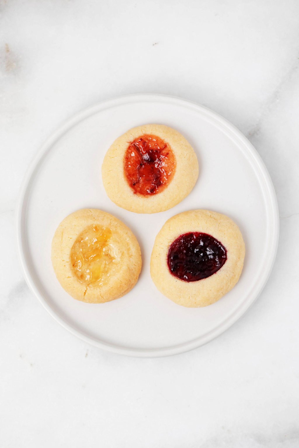 A round, rimmed dessert plate rests on a marble surface. It holds three jam filled sugar cookies.