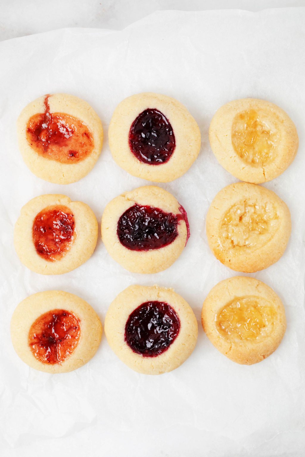 Three rows of thumbprint cookies, each filled with a different colored jam, are resting on a parchment lined baking sheet.
