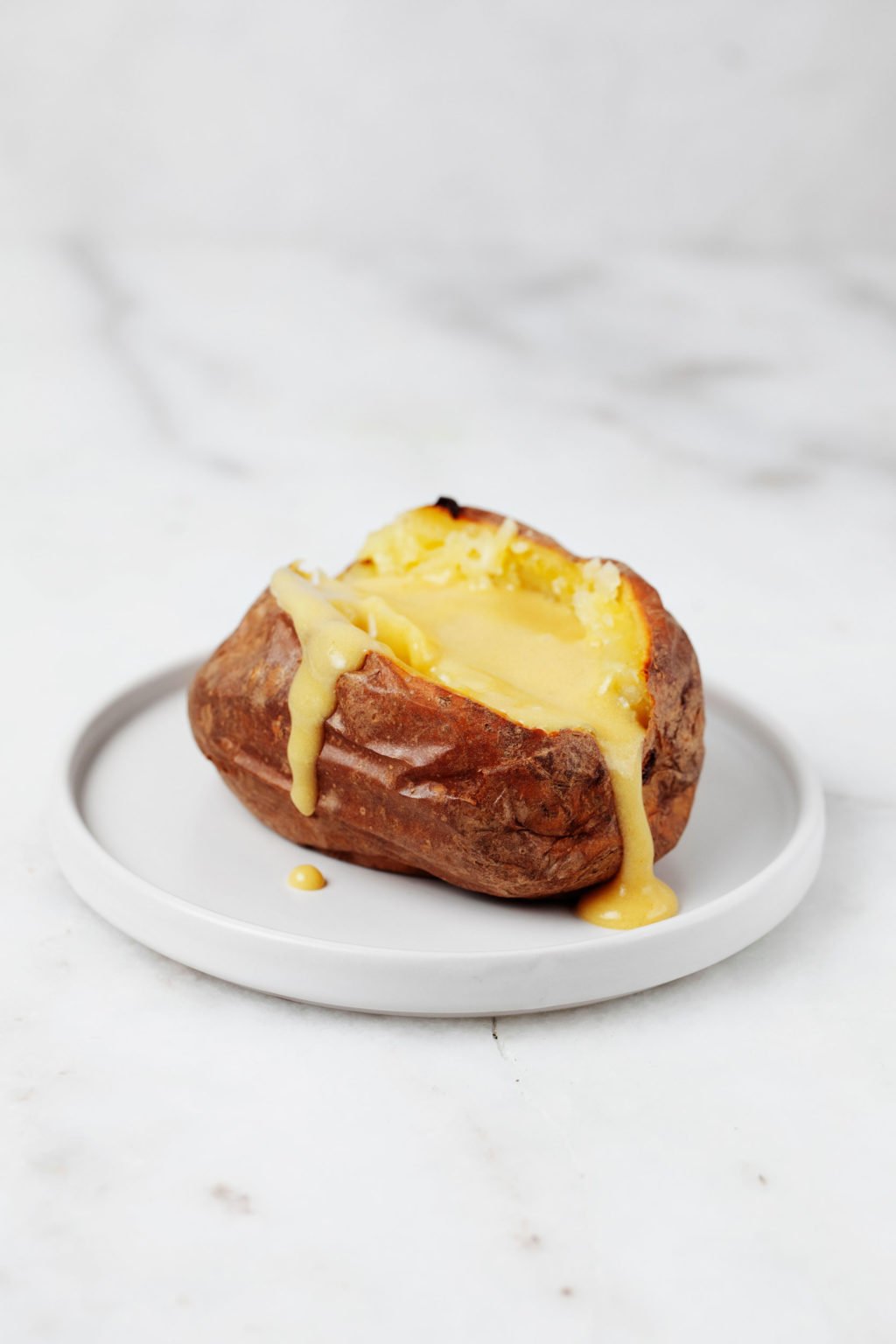 Een gepofte aardappel rust op een klein wit bordje tegen een marmeren ondergrond.  Het is bedekt met een romige vulling.
