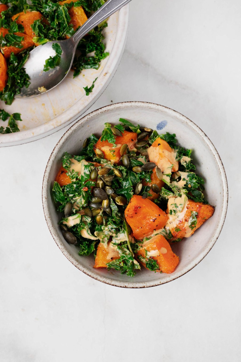 Two bowls of warm winter squash and steamed kale rest on a white marble surface.