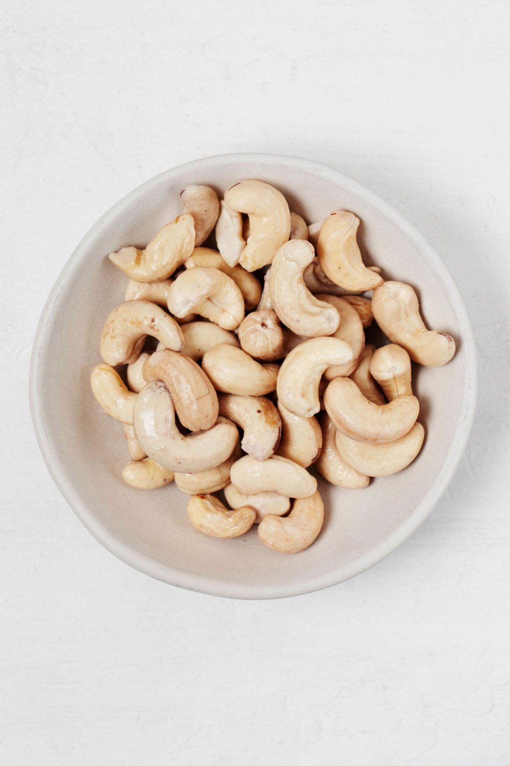 An overhead image of a white bowl, which is filled with plain cashews. It rests on a white surface.