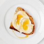 An overhead image of a round, white rimmed plate, which holds a slice of toast with vegan ricotta and agave drizzle on top.