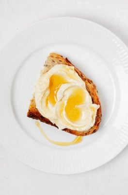 An overhead image of a round, white rimmed plate, which holds a slice of toast with vegan ricotta and agave drizzle on top.