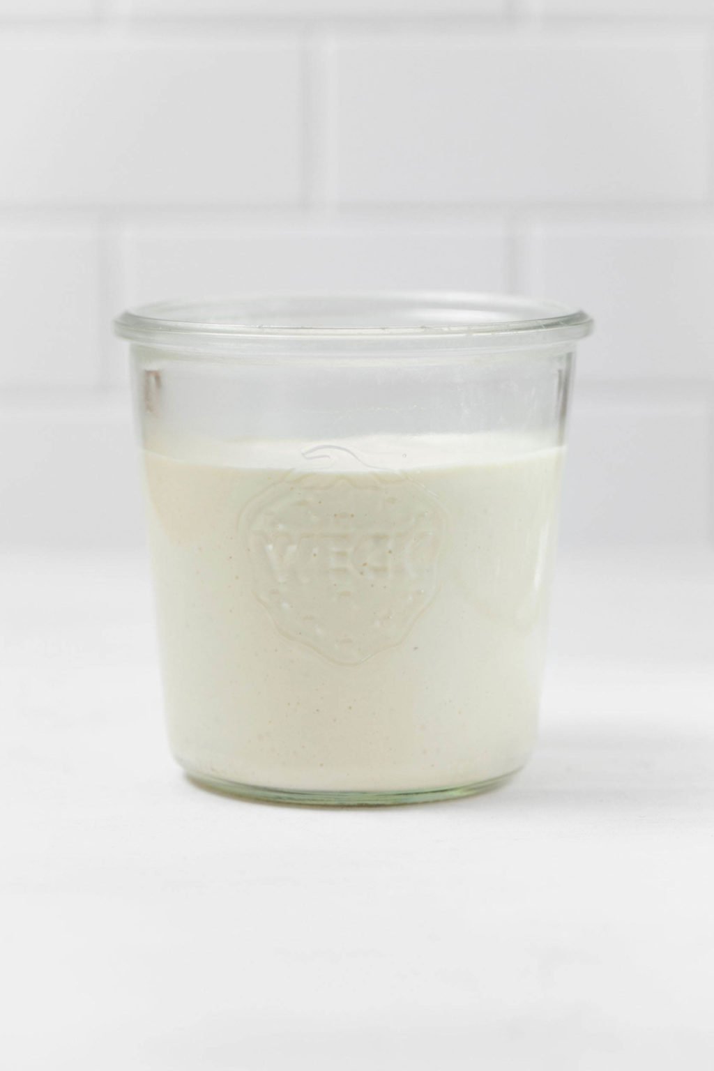 Una paleta de vidrio blanco sostiene una botella de crema agria vegana cremosa.  Se apoya sobre una superficie blanca.