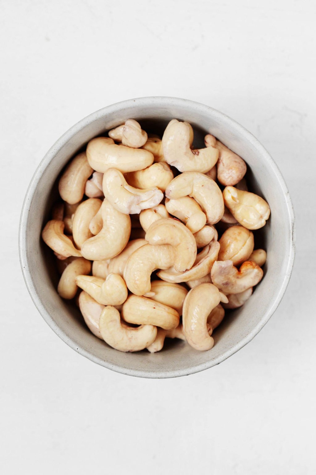 A small, white bowl rests on a white surface. It's filled with raw cashews.