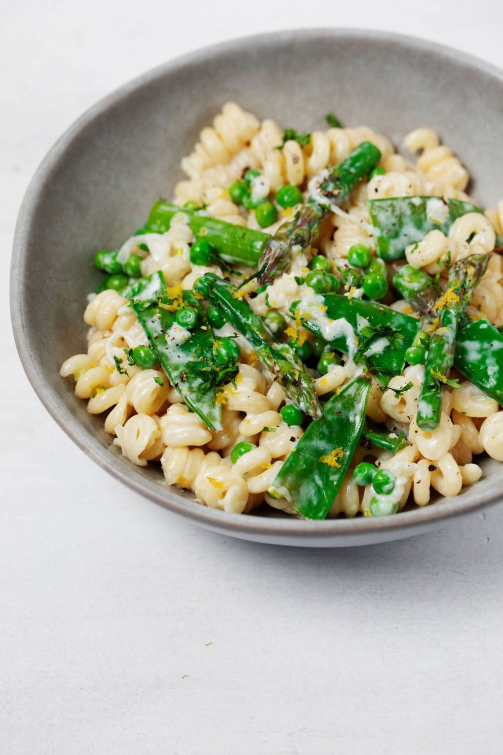 A grayish white bowl is filled with a vegan spring pasta primavera dish. It's topped with herbs and a light cream sauce.