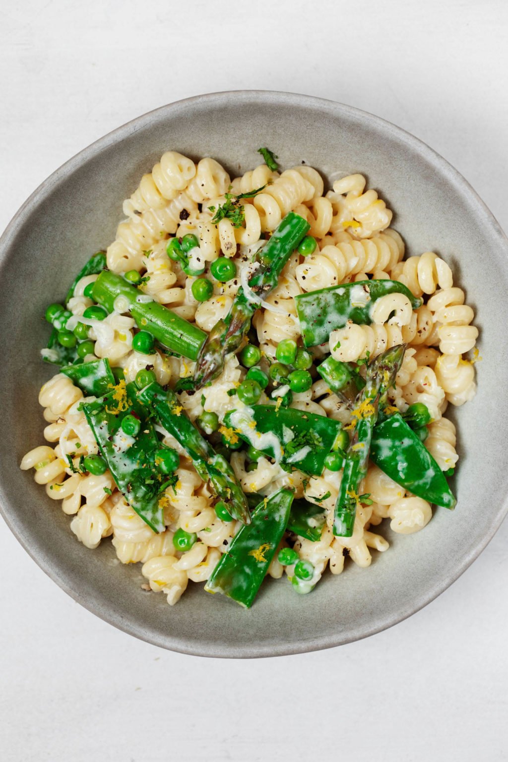 A gray and white ceramic bowl serves a vegan spring pasta primavera dish. It's topped with chopped fresh herbs and black pepper and resting on a white surface.