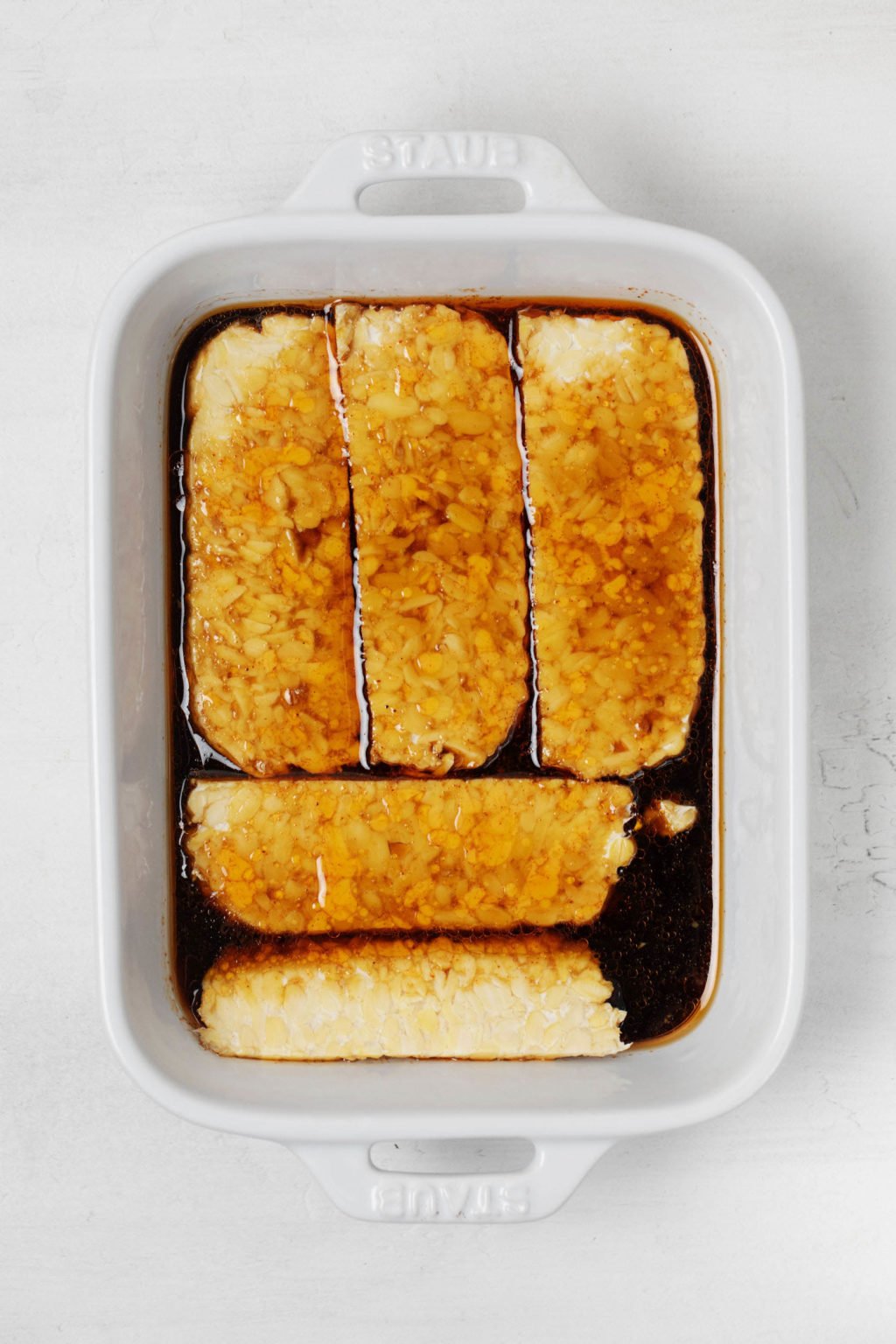 Strips of a vegan protein are lined up in a baking dish, marinating in a deep amber marinade. The dish rests on a white surface.