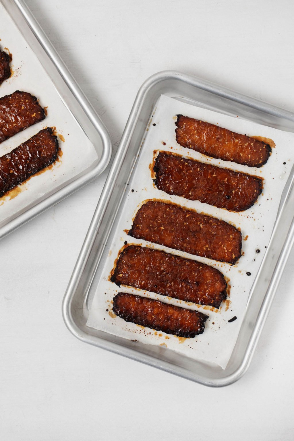 Two small baking sheets are lined with parchment paper and strips of vegan tempeh "bacon."
