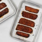 Two small baking sheets are lined with parchment paper and strips of vegan tempeh "bacon."