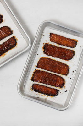 Two small baking sheets are lined with parchment paper and strips of vegan tempeh "bacon."