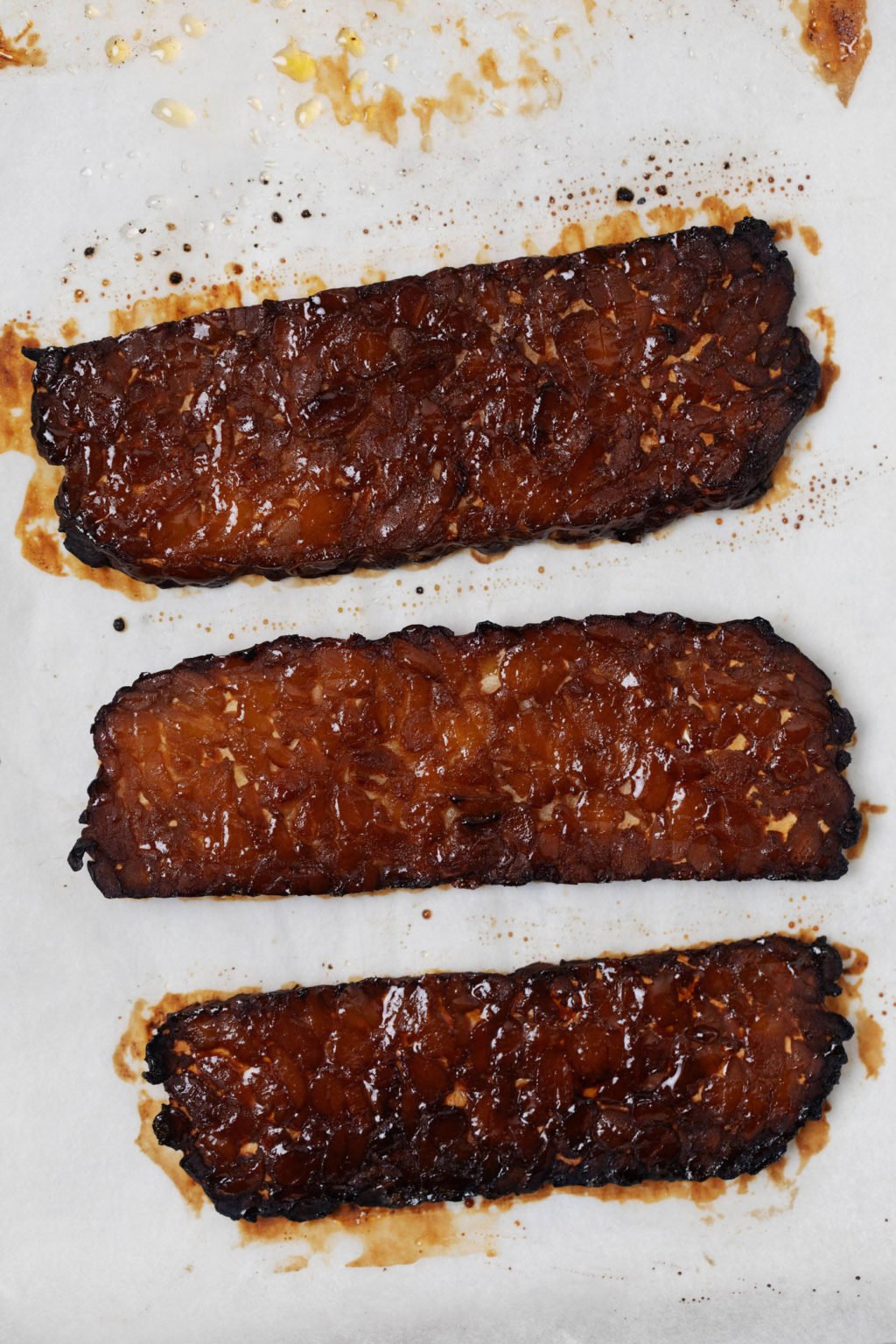 A sheet of white parchment paper has been used for baking dark brown slices of vegan tempeh "bacon."