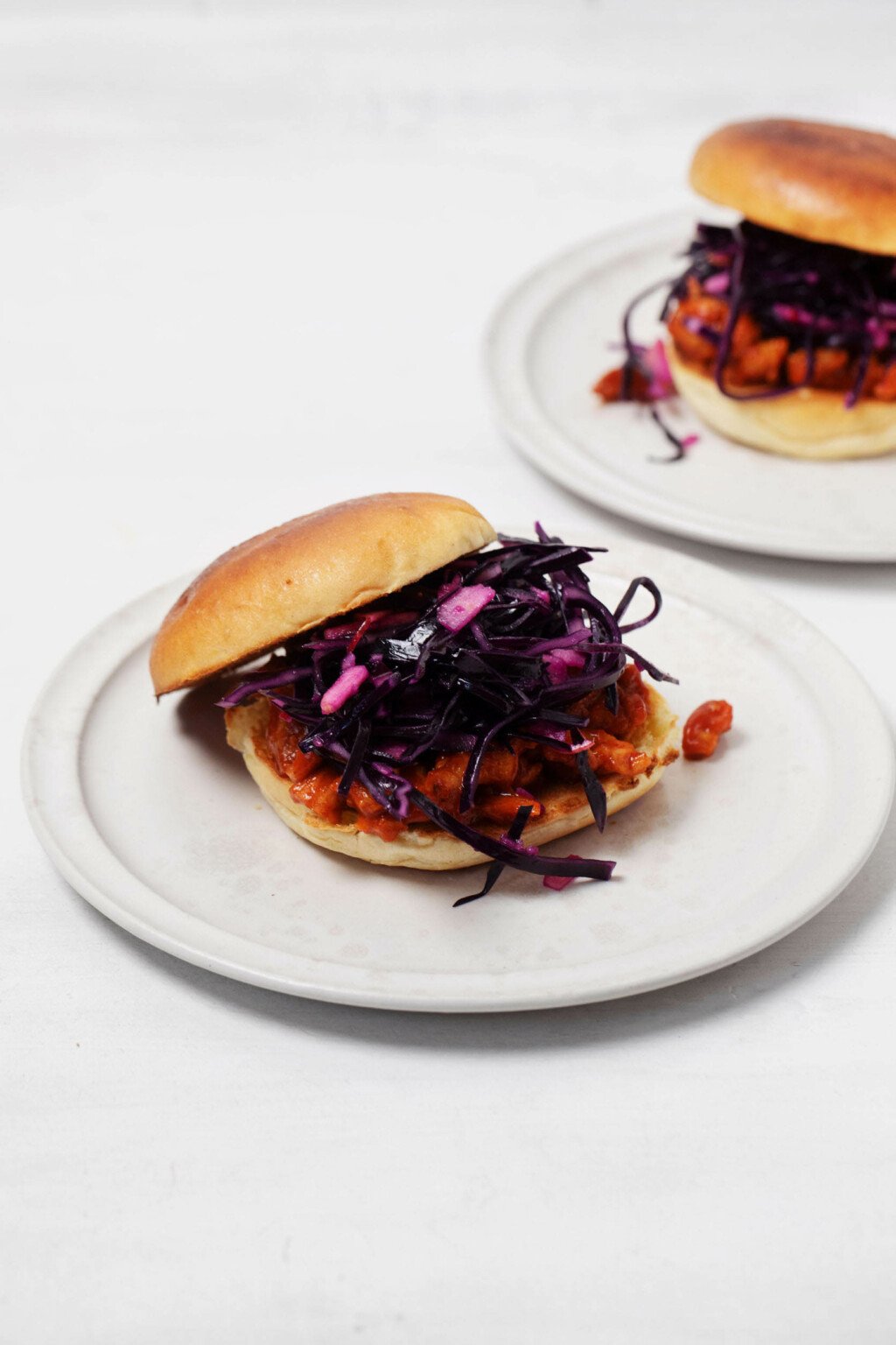 Burger buns have been stacked with a vegan version of BBQ chicken and a crunchy red cabbage slaw. They rest on two round, rimmed white plates.