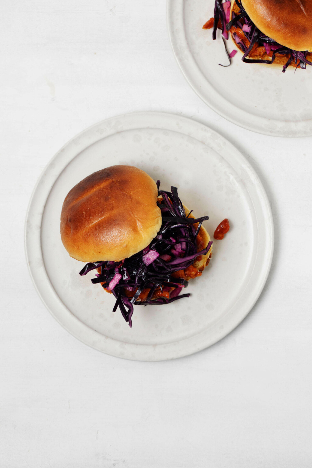 A toasted burger bun is stacked with a red cabbage slaw and a vegan protein. It rests on a round, white plate.