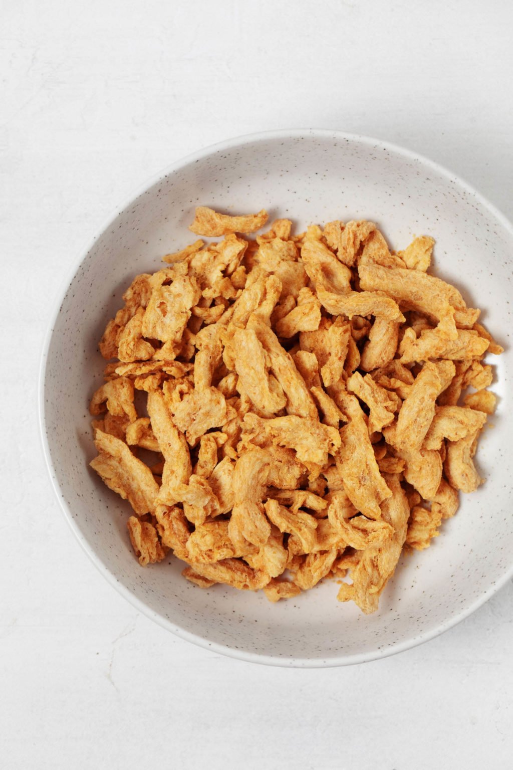 A bowl has been filled with soy curls. The bowl rests on a white surface.