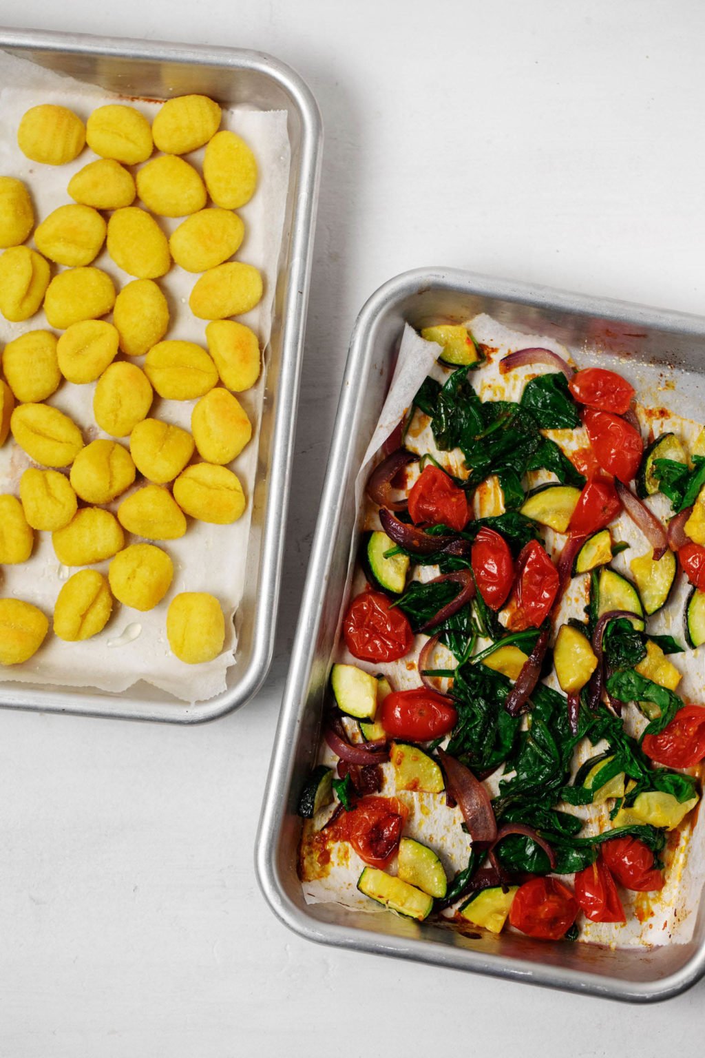 Two sheet pans have been covered with roasted vegetables and crispy gnocchi. They rest on a white surface.