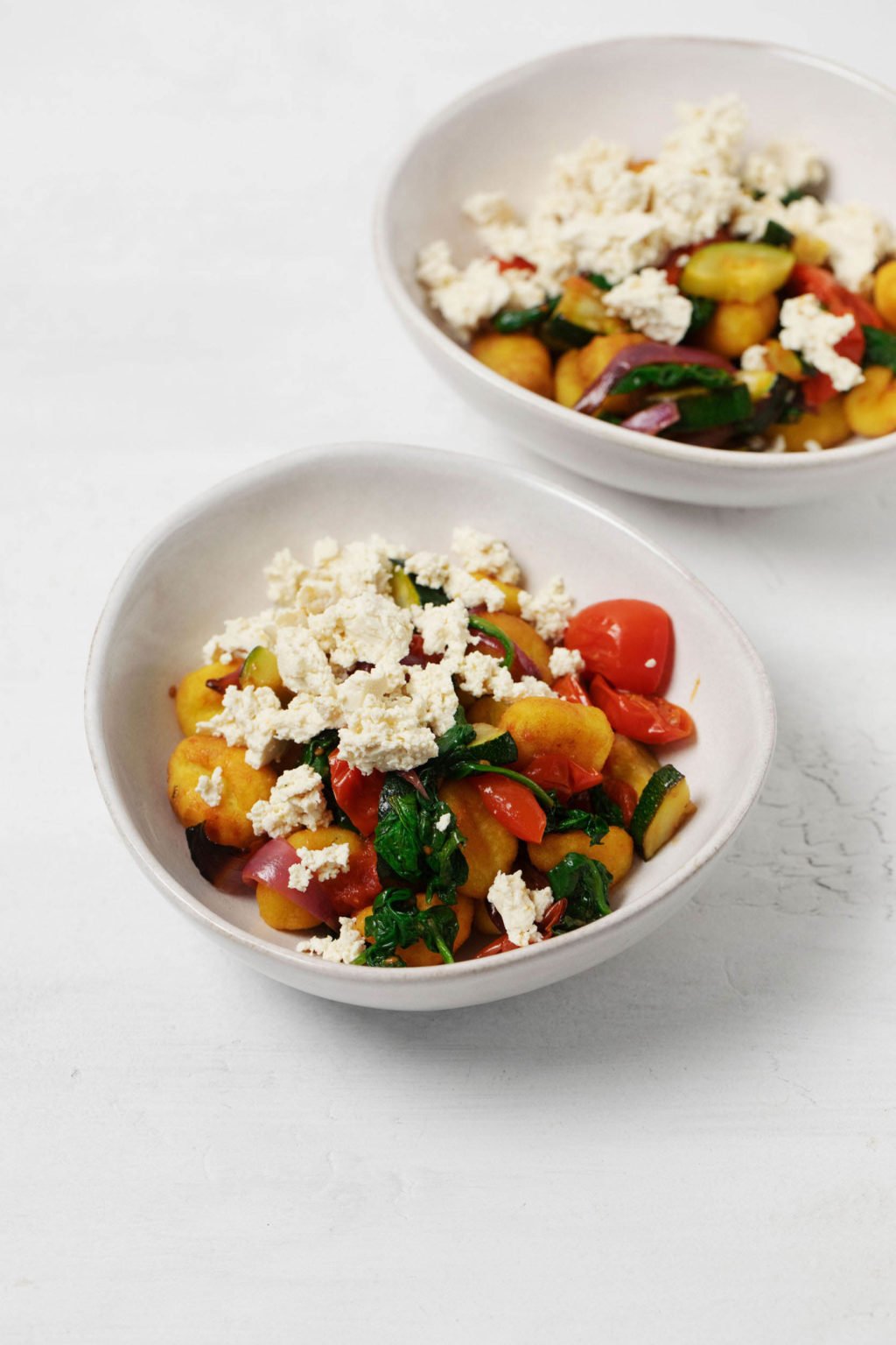 Two white bowls have been filled with a vegan sheet pan gnocchi meal. They're topped with crumbled vegan feta cheese.