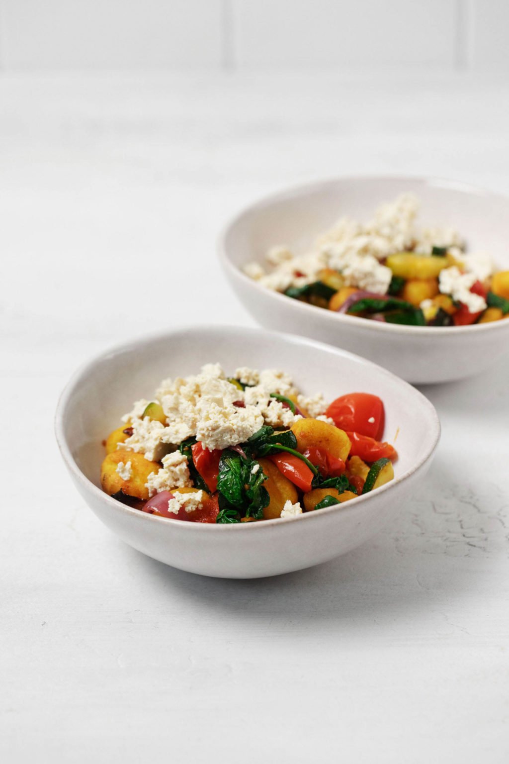 Two white, asymmetrical round bowls are serving a vegan sheet pan gnocchi meal. They're topped with crumbles of a homemade, dairy-free cheese.