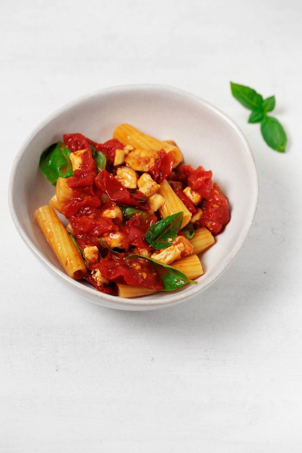 A white bowl of pasta with tomatoes and torn fresh basil leaves. It rests on a white surface.
