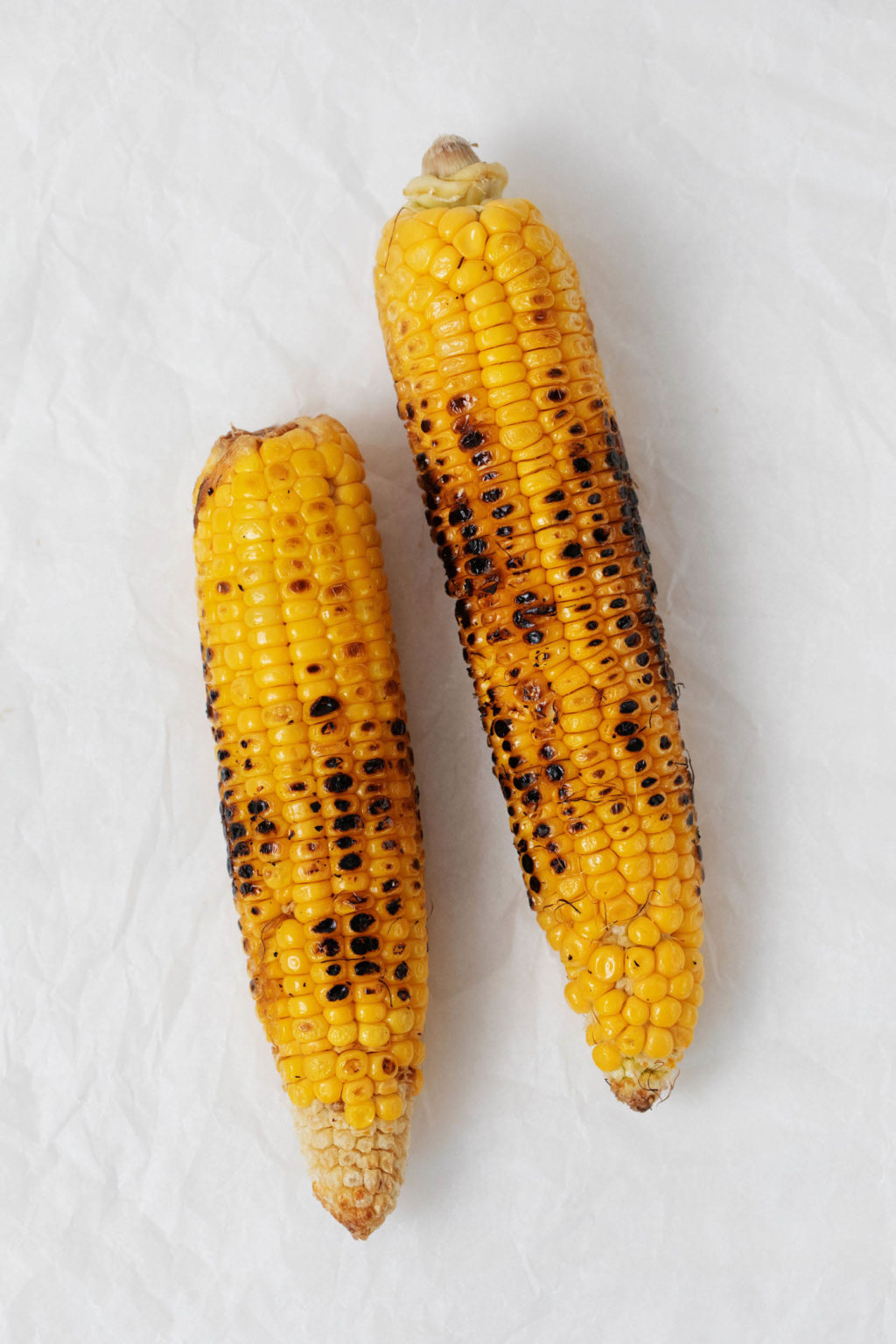 Two ears of grilled corn are resting on a sheet of parchment paper.