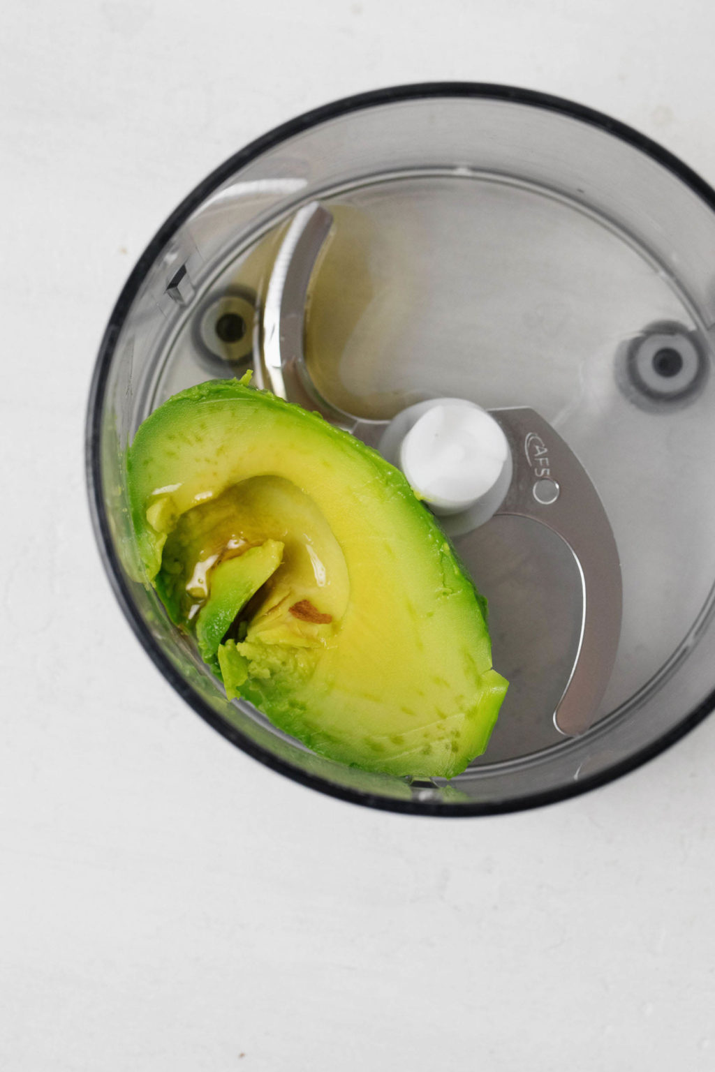 An avocado is in the bowl of a food processor, which has been fitted with the S blade.