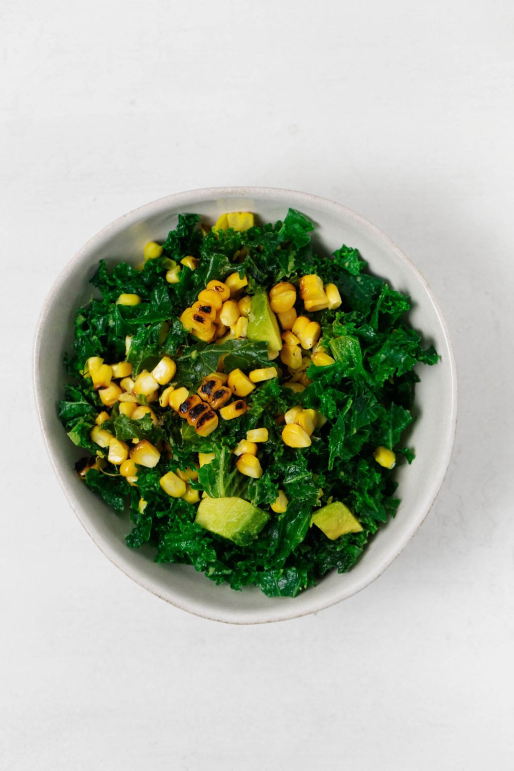 A vegan grilled corn avocado salad with kale has been served in a white ceramic bowl.