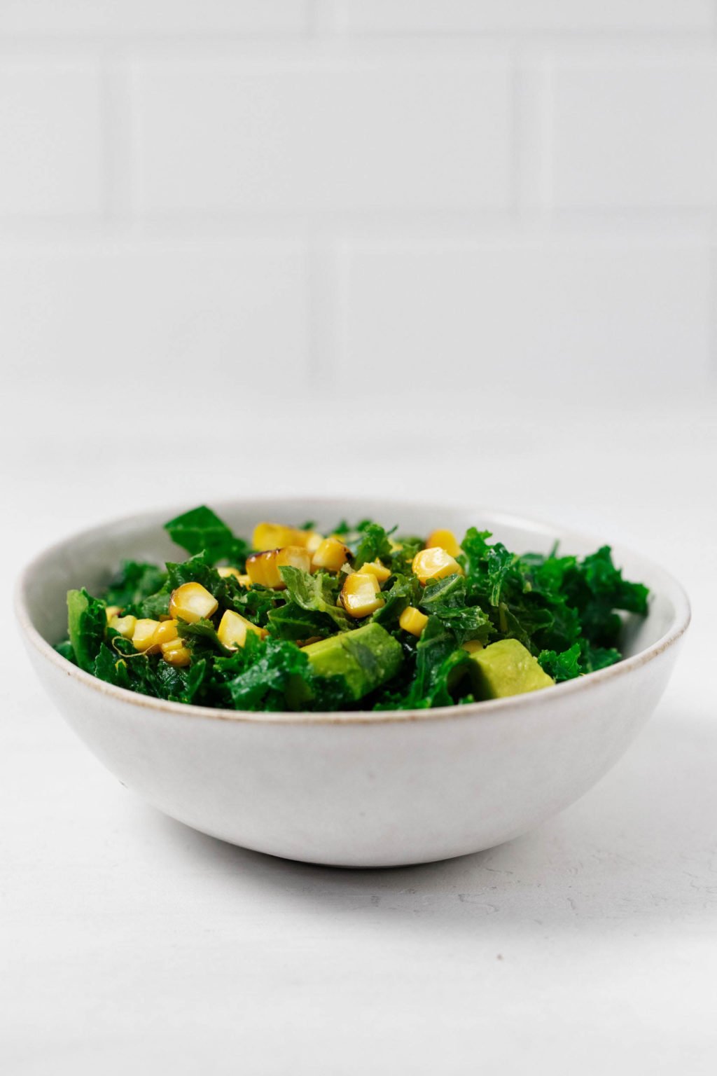 A bowl of bright green kale and other vegetables rests on a white surface, with a white tiled wall in the background.