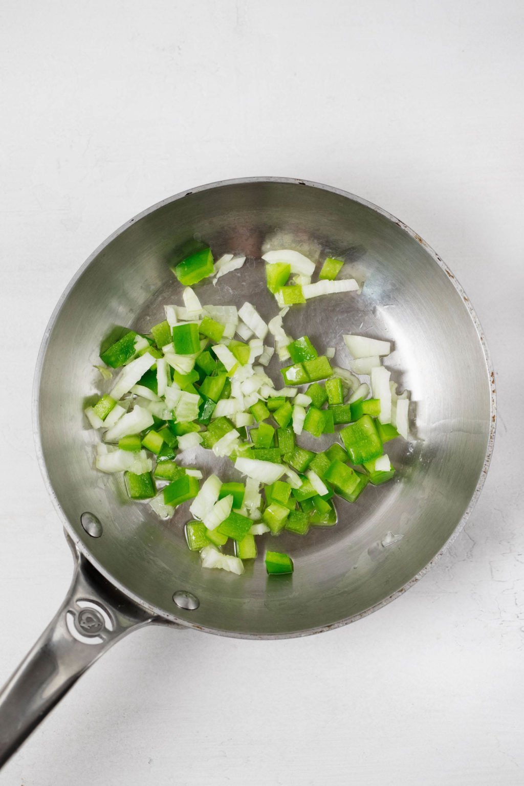 Chopped onion and green bell pepper are being sautéed in a small frying pan. It rests on a white surface.