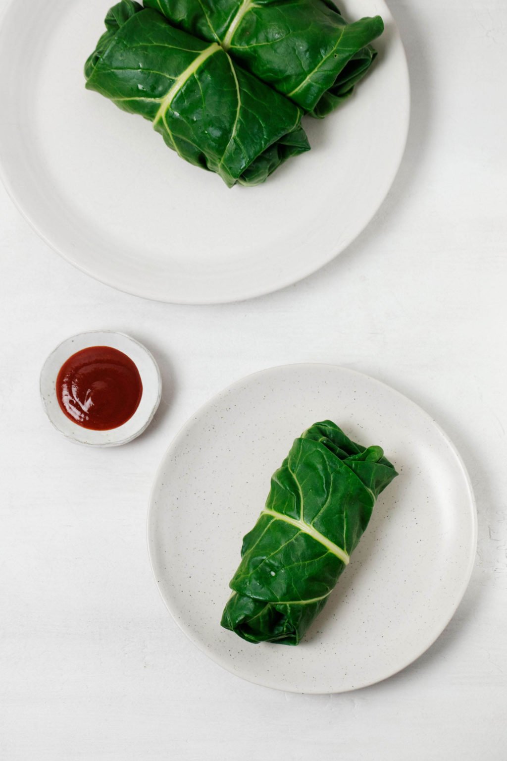 Leafy greens have been stuffed with whole grains and legumes. They're served on small white plates with barbecue sauce nearby.