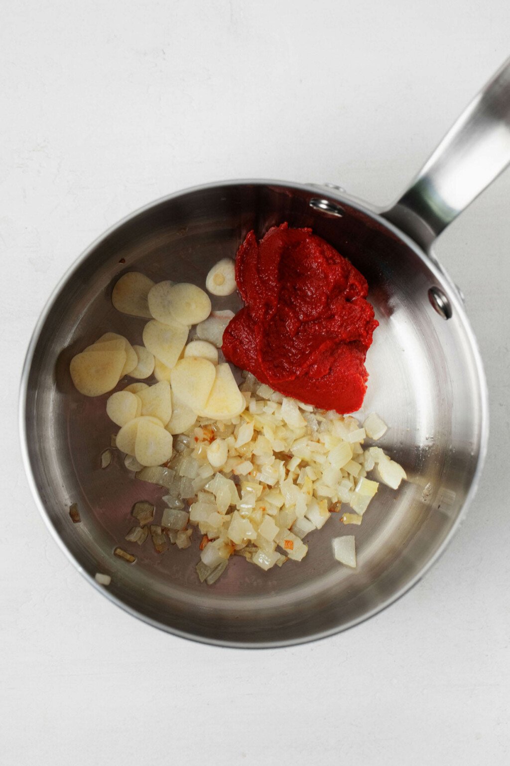 Garlic, onions, and tomato paste are being simmered in a silver, stainless steel sauce pot.