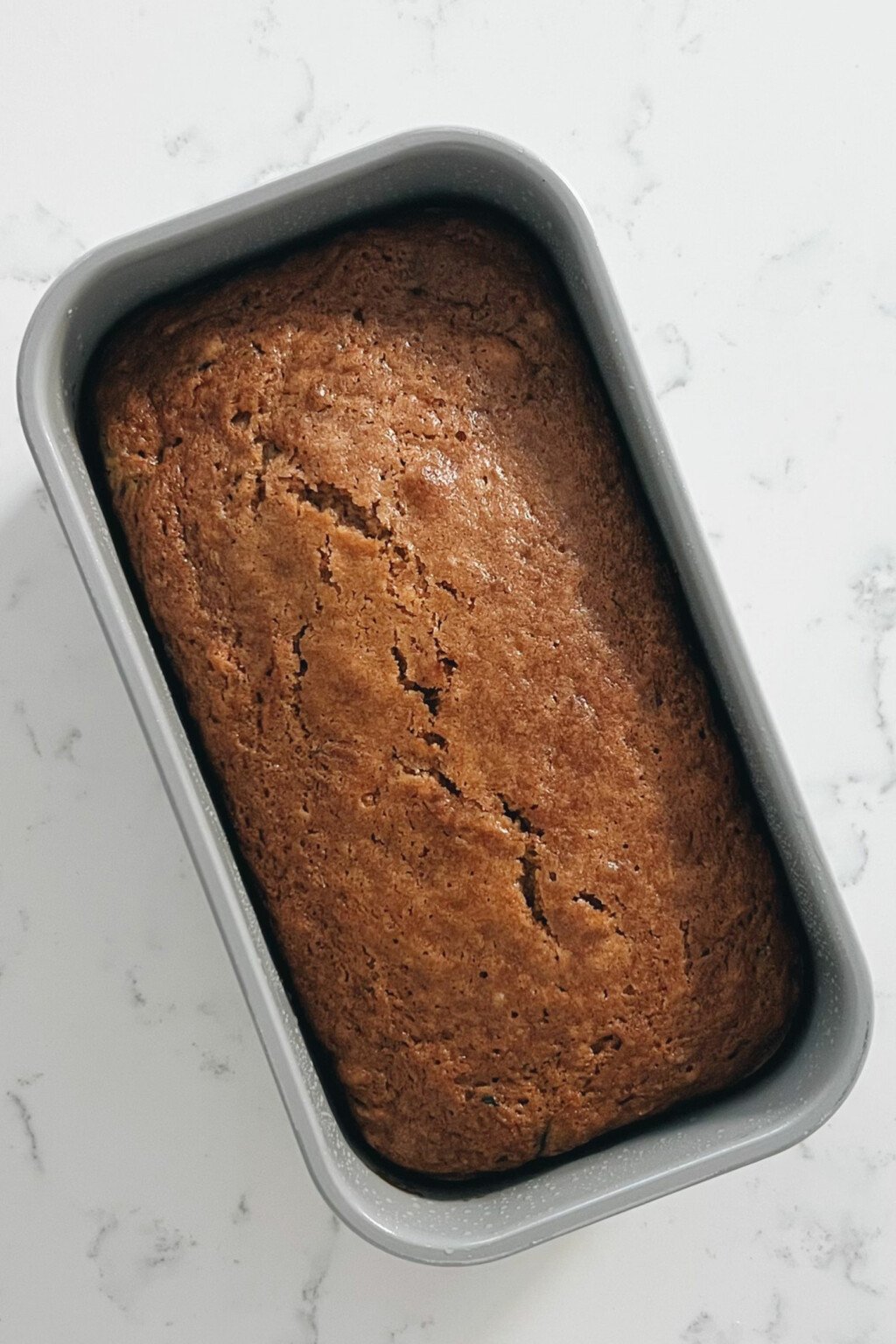 A gray, rectangular loaf pan rests on a white marbled surface. It holds freshly baked bread.